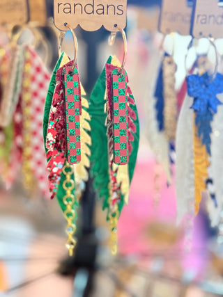 Festive tiered earrings featuring red and green glitter checkerboard pieces layered over green, gold, and floral faux leather feathers, with gold hoops and chain fringe. Displayed on a "randans" card.