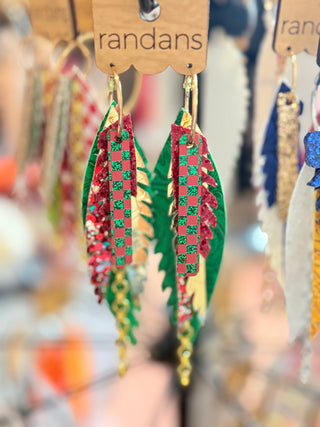 Festive tiered earrings featuring red and green glitter checkerboard pieces layered over green, gold, and floral faux leather feathers, with gold hoops and chain fringe. Displayed on a "randans" card.