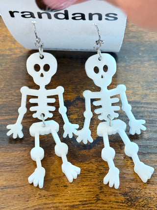 White dangling skeleton earrings made from lightweight acrylic, each with arms raised showing the middle finger. Displayed on a “randans” card on a wooden background.