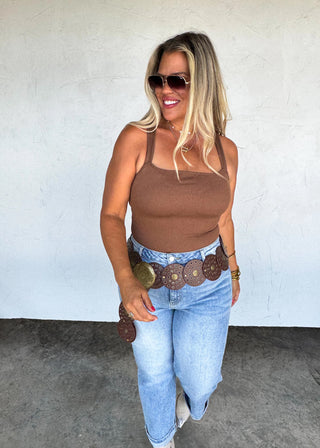 Woman wearing a brown tank top and blue jeans with a conch belt, standing against a white wall.
