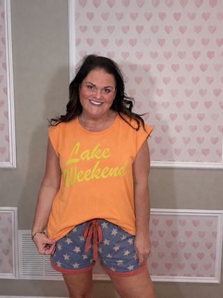 Woman wearing an orange 'Lake Weekend' shirt and star-patterned shorts in a room with heart-shaped wallpaper.