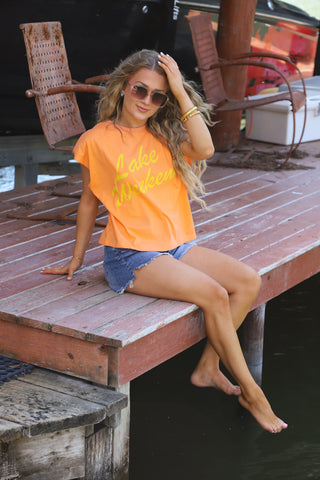 Woman sitting on a wooden dock wearing an orange t-shirt with text and denim shorts.