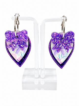 A pair of dangling earrings displayed on a clear acrylic stand against a white background. Each earring features a glittery purple heart-shaped dangle with a guitar-pick pattern in purple, pink, and white, attached to silver-tone hoops.