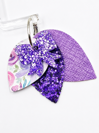 A close-up of earrings with three overlapping leaf-shaped pieces: one floral print, one purple glitter bow, and one purple textured fabric, attached to a silver metal ring.