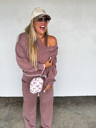 Woman wearing a mauve off-shoulder top and pants with a cap and sunglasses, holding a patterned bag against a white background.