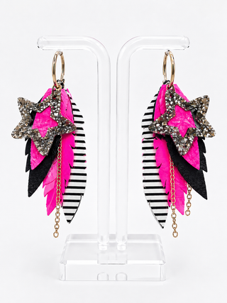 A pair of long, layered dangle earrings displayed on a clear acrylic stand against a white background. The earrings feature bright hot pink feathers, black feathers with a black-and-white striped pattern, glittery silver star charms, and gold-tone rings and chains.