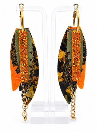 A pair of long dangling earrings displayed on clear acrylic stands against a white background. Each earring has a gold hoop and chain, with layered feather-like pieces in orange, olive green, black, and brown camouflage, and a glittery orange vertical stripe.