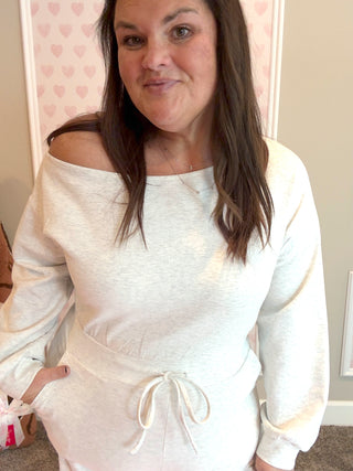 Woman wearing an ash gray off-shoulder jumpsuit in a room with pink heart wallpaper.