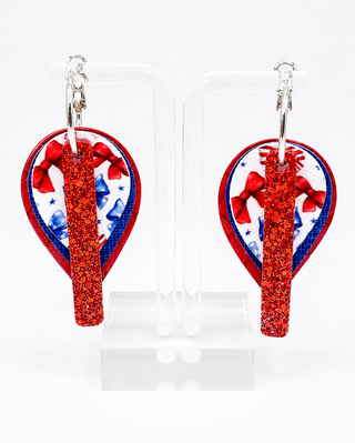 A pair of patriotic-themed dangle earrings displayed on clear acrylic stands against a white background. The earrings feature a glittery red vertical bar, a red, white, and blue patterned heart-shaped background, and silver-tone hooks.