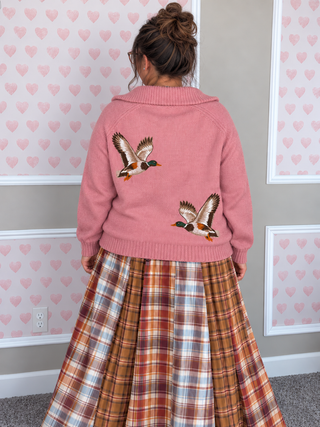A person viewed from behind wearing a pink knit cardigan with two embroidered flying ducks on the back, paired with a brown, orange, and white plaid skirt. The background features a gray wall with white trim and pink heart-patterned wallpaper.