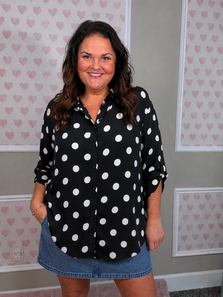 A woman wearing a black button-down blouse with white polka dots and a denim mini skirt, standing in front of a wall with pink heart-patterned wallpaper.