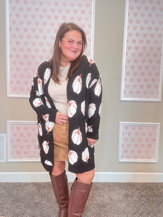 Woman wearing a black cardigan with white pumpkins, beige skirt, and brown knee-high boots in a room with heart-patterned wallpaper.