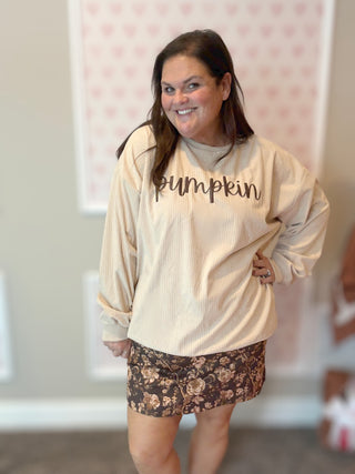 Woman wearing a beige sweatshirt with 'Pumpkin' text and floral shorts indoors.