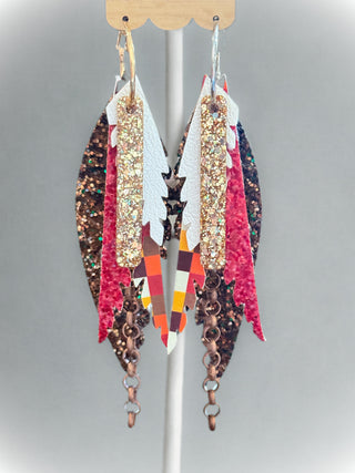 Pair of long layered faux leather earrings with gold glitter, orange-red plaid pattern, white and red textured accents, and dangling bronze chains. Displayed on a rotating rack with a “randans” earring card.
