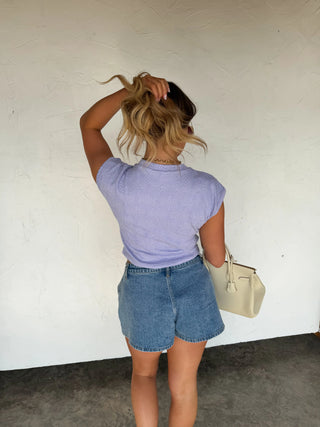 Person wearing a purple shirt and denim shorts, holding a beige handbag against a white wall.
