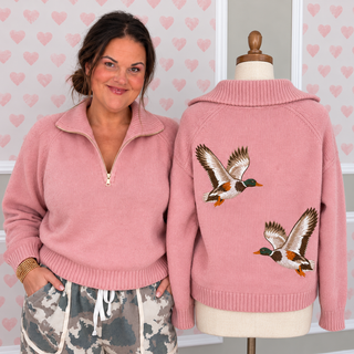 A woman wearing a pink half-zip sweater stands beside a mannequin displaying the back of the same sweater. The sweater has two embroidered mallard ducks on the back.
