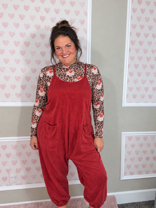A woman wearing a red overall jumpsuit over a leopard print long-sleeve shirt, standing in front of a wall with pink heart-patterned wallpaper.