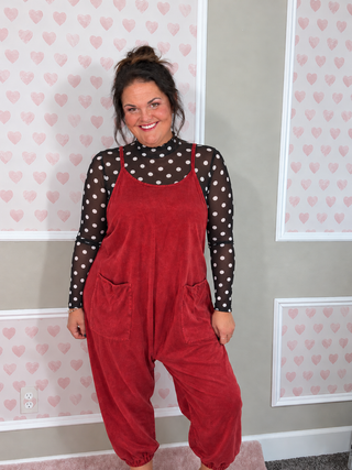 A woman wearing a red hooded jumpsuit over a black and white polka dot long-sleeve top, standing in front of a wall with pink heart wallpaper.