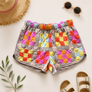 Flat lay of colorful quilted patchwork shorts with neutral sandals, sunglasses, and straw hat styling.

