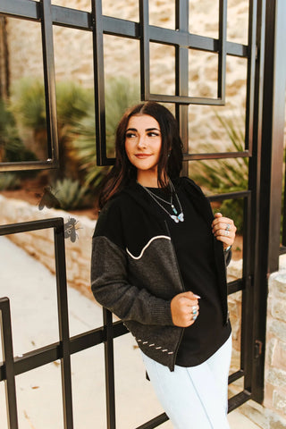 Woman wearing a black and gray sweater with white pants standing in front of a metal gate.