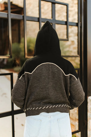 Person wearing a black and gray hoodie with white stitching, standing in front of a glass door.