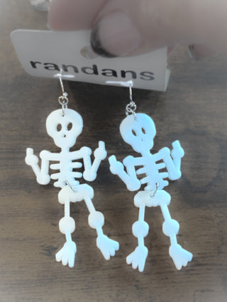 White dangling acrylic skeleton earrings with expressive arm positions and cute eye holes. Held on a “randans” earring card over a wooden table.