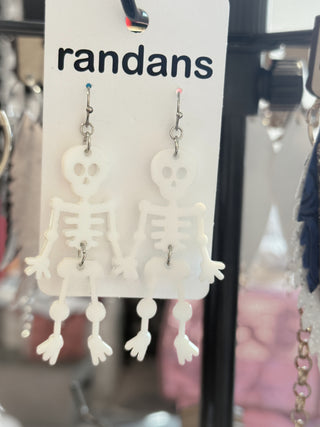 White dangling skeleton earrings made from lightweight acrylic, each with arms raised showing the middle finger. Displayed on a “randans” card on a wooden background.