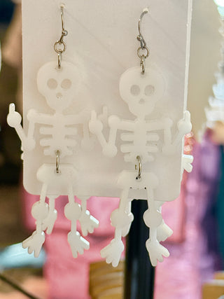 White dangling acrylic skeleton earrings with expressive arm positions and cute eye holes. Held on a “randans” earring card over a wooden table.