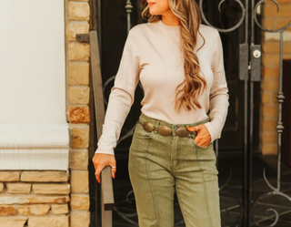 Woman wearing a beige sweater and green pants standing in front of a stone wall.