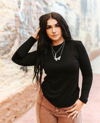 Woman wearing a black long-sleeve top and brown pants with a blurred natural background