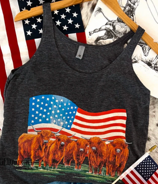 A dark heather gray racerback tank top hanging on a wooden hanger, featuring a graphic print of Highland cows in front of a waving American flag. The tank top is displayed against a background with another American flag and a western-themed print.