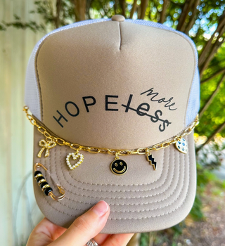 Cream foam trucker hat with Hope More graphic, gold hat chain, decorative pin, and adjustable mesh back.