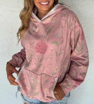A woman wearing a dusty pink floral print hoodie with white lace trim on the hood and pocket edges, paired with light blue denim shorts, posing against a white textured wall.