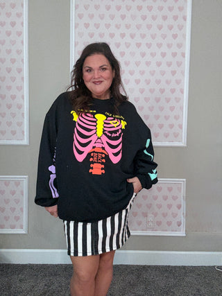 Person wearing a black sweatshirt with a colorful skeleton design and a striped skirt in front of a wall with heart patterns.