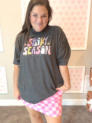 Person wearing a dark gray t-shirt with 'Spooky Season' text and a pink checkered skirt in a room with heart-patterned wallpaper.