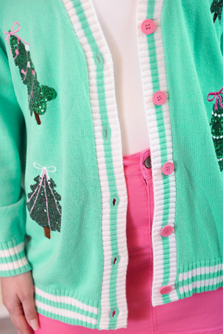 Green cardigan with festive tree designs and pink buttons on a person wearing pink pants.