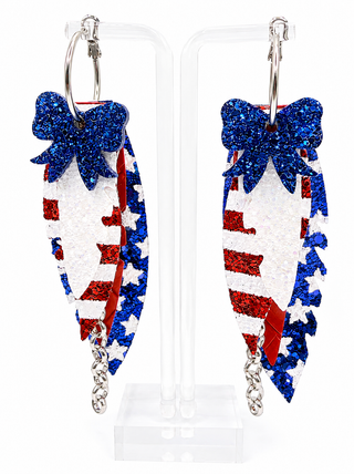 A pair of patriotic hoop earrings displayed on a clear acrylic stand. The earrings feature silver-tone hoops and dangling glittery shapes with blue bows, red and white sequins, and a small silver chain dangle.
