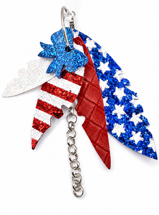 A patriotic-themed earrings featuring three stylized feathers in red, white, and blue glitter and textured finishes, attached to a silver metal ring and chain.