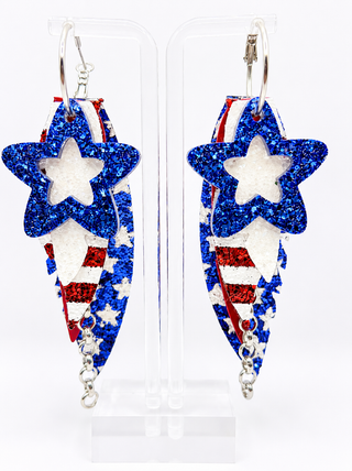 A pair of patriotic dangle earrings displayed on clear acrylic stands against a white background. The earrings feature a glittery blue star on the top, a white star in the center, and a red, white, and blue glittery American flag pattern underneath with small silver chain drops.