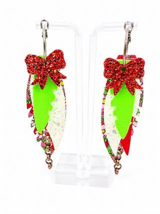 A pair of dangling earrings displayed on clear acrylic stands against a white background. Each earring has a bright red glitter bow at the top, a lime green textured side panel, a white glitter center panel, and a multicolor patterned ruffle along the bottom edge.