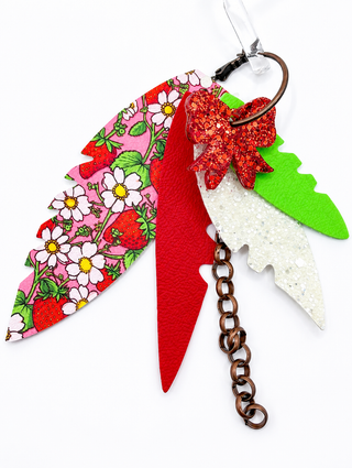 A set of colorful, leaf-shaped earrings made from different materials, including a pink strawberry print leather, a red faux-leather piece, a glittery red bow, a green glitter piece, and a white glitter piece, attached to a metal key ring and chain.