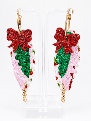 A pair of gold hoop earrings with long, leaf-shaped charms decorated with red glitter bows, green glitter holly leaves, and pink glitter sections with a strawberry-and-white pattern, displayed on clear acrylic stands against a white background.