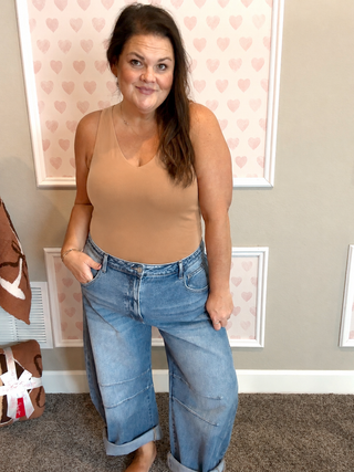 Woman wearing a tan seamless v-neck bodysuit paired with relaxed light wash cuffed jeans in a cozy feminine boutique setting with soft pink heart wall décor.