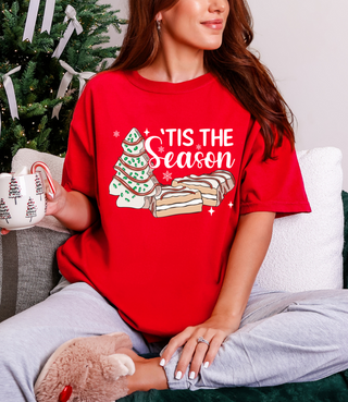 Person wearing a red 'Tis the Season t-shirt with a Christmas tree and cookies design, holding a mug.