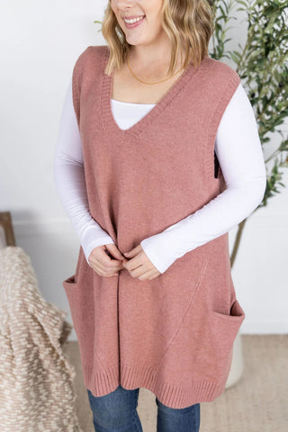 Woman wearing rose tunic sweater vest over a white long sleeve top, styled with black leggings and boots.