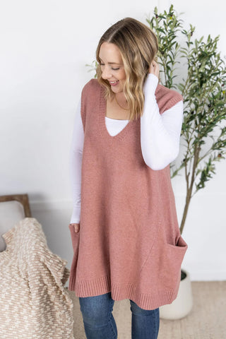 Woman wearing rose tunic sweater vest over a white long sleeve top, styled with black leggings and boots.