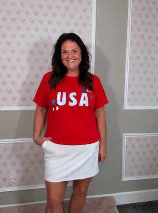 A woman wearing a red short-sleeve t-shirt with 'USA' printed in white letters with blue stars, paired with a white mini skirt, standing in front of a gray wall with pink heart wallpaper.