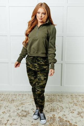 Woman wearing a green tracksuit with camouflage pants against a white tiled wall.