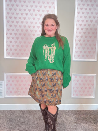 Person wearing a green sweater with a graphic design and a western patterned skirt, standing in front of a wall with heart designs.