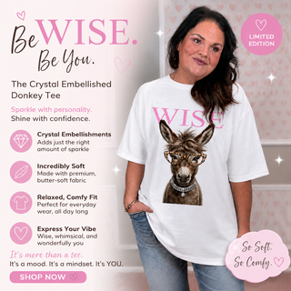 A woman wearing a white graphic t-shirt with 'WISE' and a donkey illustration, set against a pink promotional background with text highlighting the shirt's features and crystal embellishments.